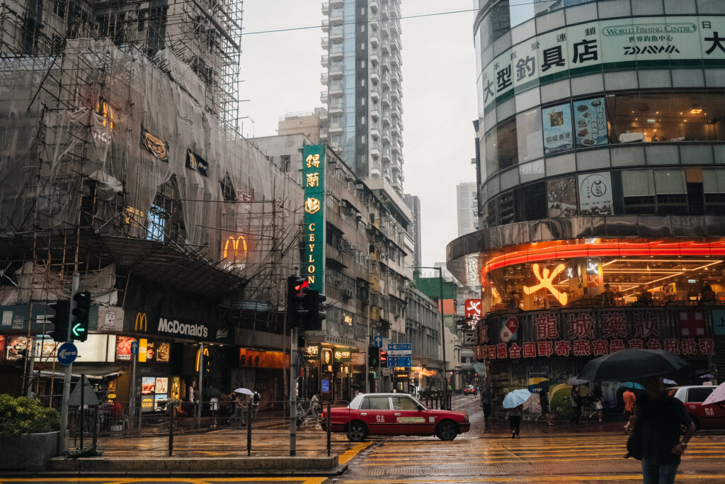 High-angle view of a bustling city street in Hong Kong during rainy weather, showcasing neon signs, tall buildings, and pedestrians with umbrellas, capturing the vibrant urban atmosphere for travel and tourism enthusiasts.